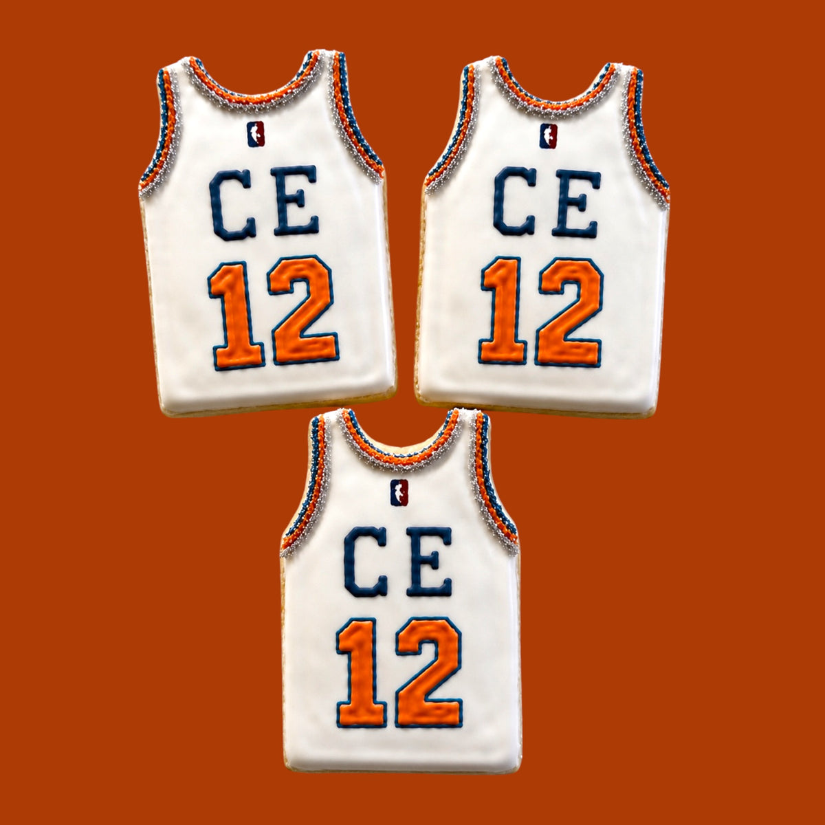Three custom Knicks basketball jersey cookies in white with blue CE letters, orange number 12, NBA logo, and beaded trim on orange background
