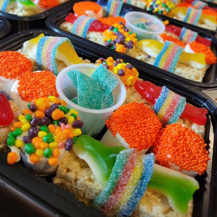 Colorful candy sushi pieces arranged in black takeout containers with rainbow gummy belts, orange candy roe, sour gummy candy, green apple slices, and small candy cup with butterfly decoration