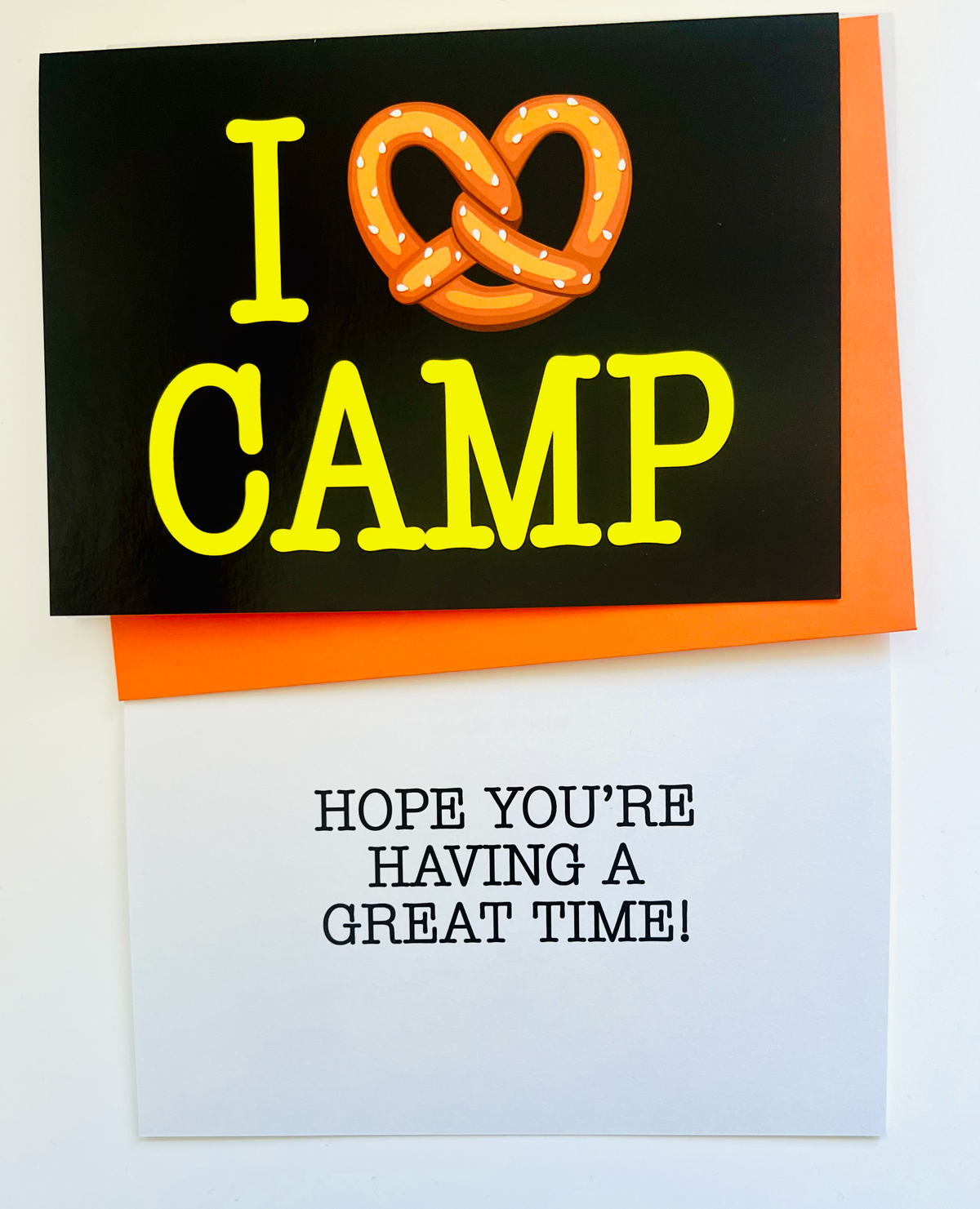 I Pretzel Camp greeting card with black background and yellow text featuring an illustrated orange pretzel with salt, orange envelope visible in background
