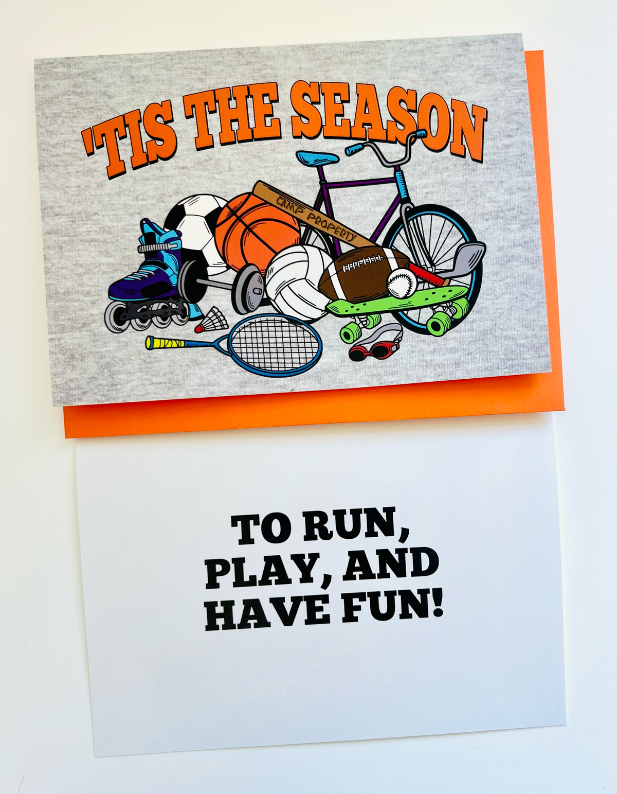 'Tis the Season camp greeting card featuring colorful illustration of sports equipment including soccer ball, basketball, football, bicycle, rollerblades, tennis racket and skateboard on heather gray background with orange text, shown with orange envelope