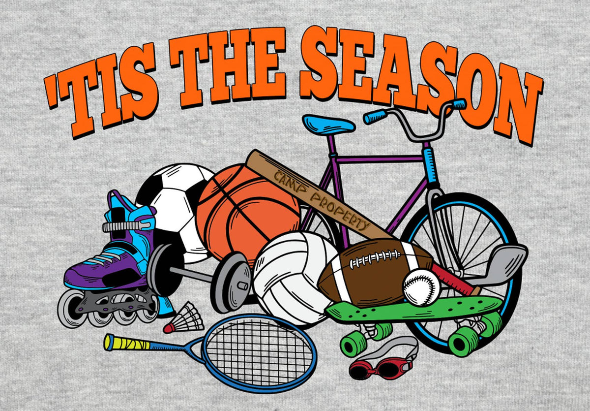 Close-up of 'Tis the Season camp greeting card on heather gray background showing detailed sports equipment illustration with soccer ball, basketball, football, bicycle, tennis racket, skateboard, rollerblades and goggles beneath orange lettering