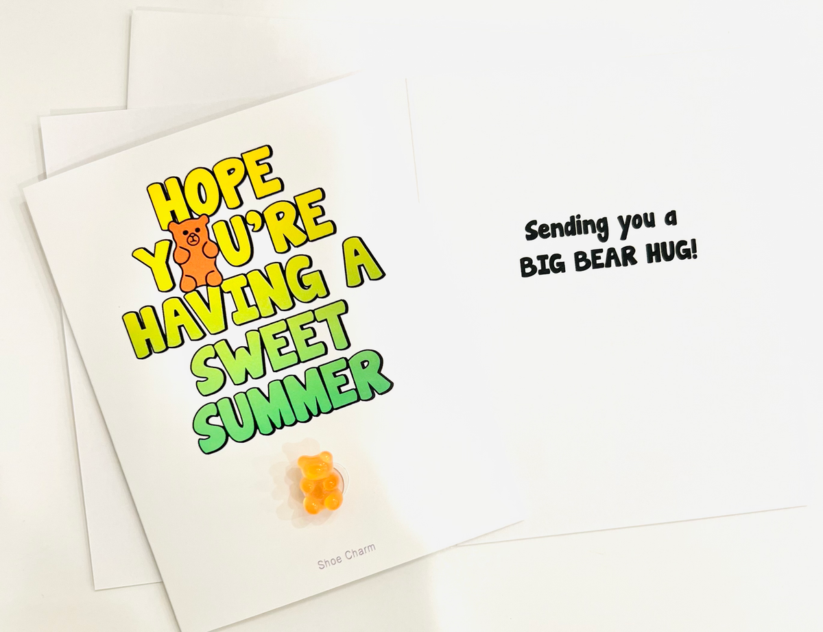 Sweet summer camp greeting card with colorful yellow and green "Hope You're Having A Sweet Summer" text, featuring attached orange gummy bear shoe charm, with text "Sending you a BIG BEAR HUG" on white card