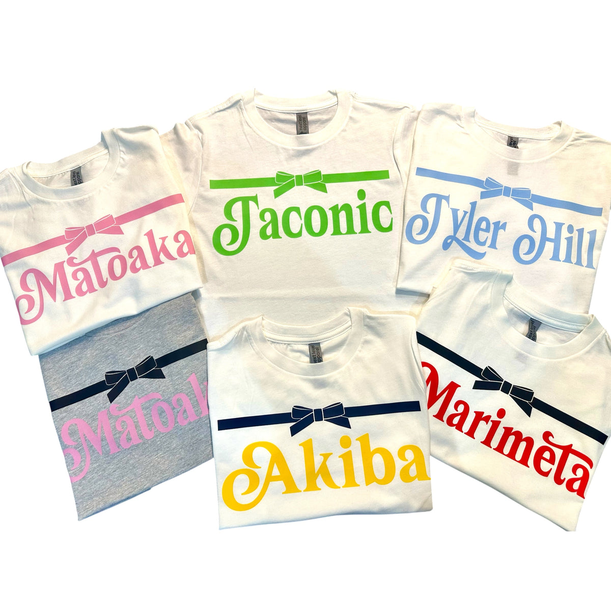 Collection of personalized ribbon camp tees including Maloaka in pink, Taconic in green, Tyler Hill in blue, Akiba in yellow, and Marimeta in red with ribbon bow detail across chest