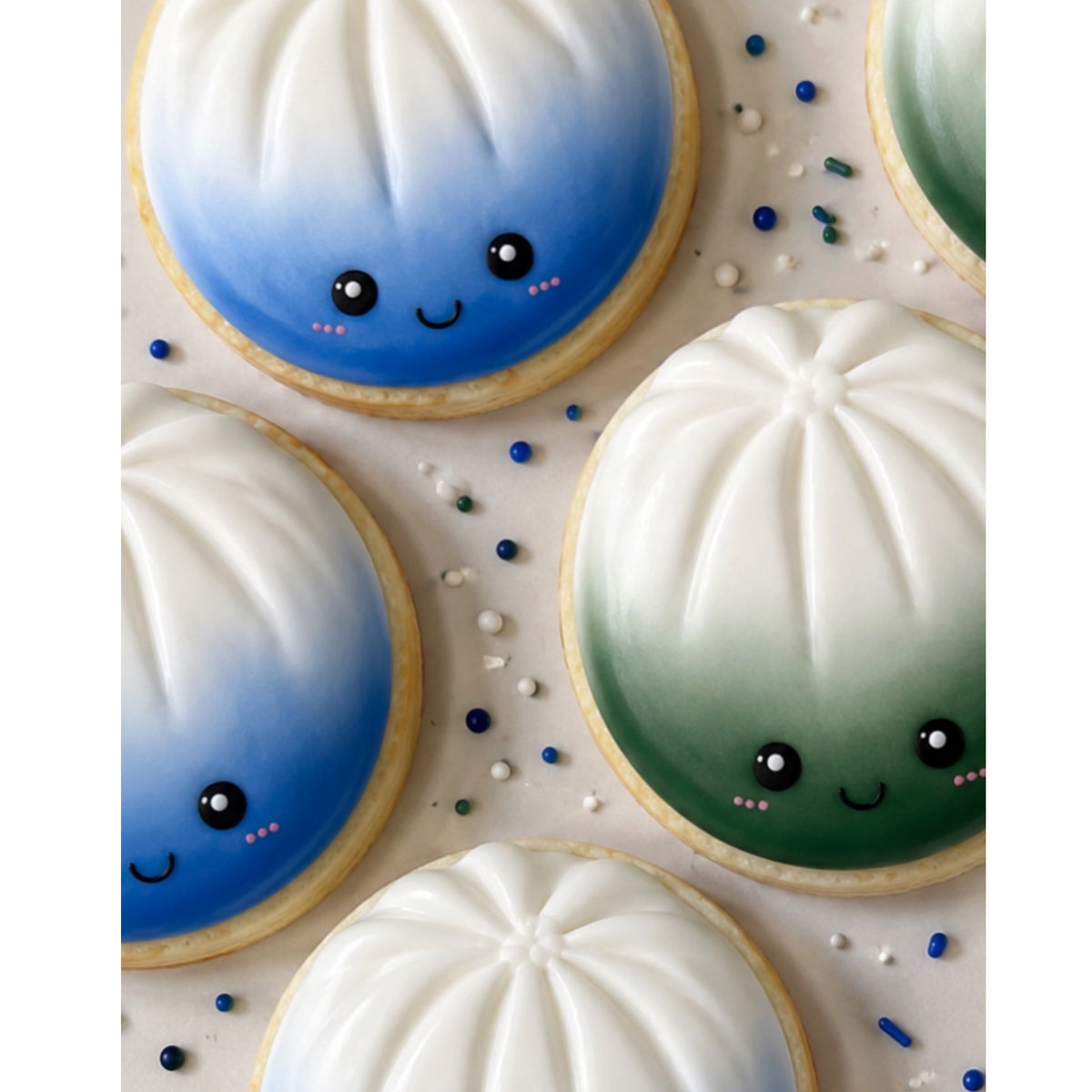 Sweet Dumplings custom cookies with white ridged tops and kawaii faces in blue and green ombre frosting, arranged on white surface with decorative sprinkles