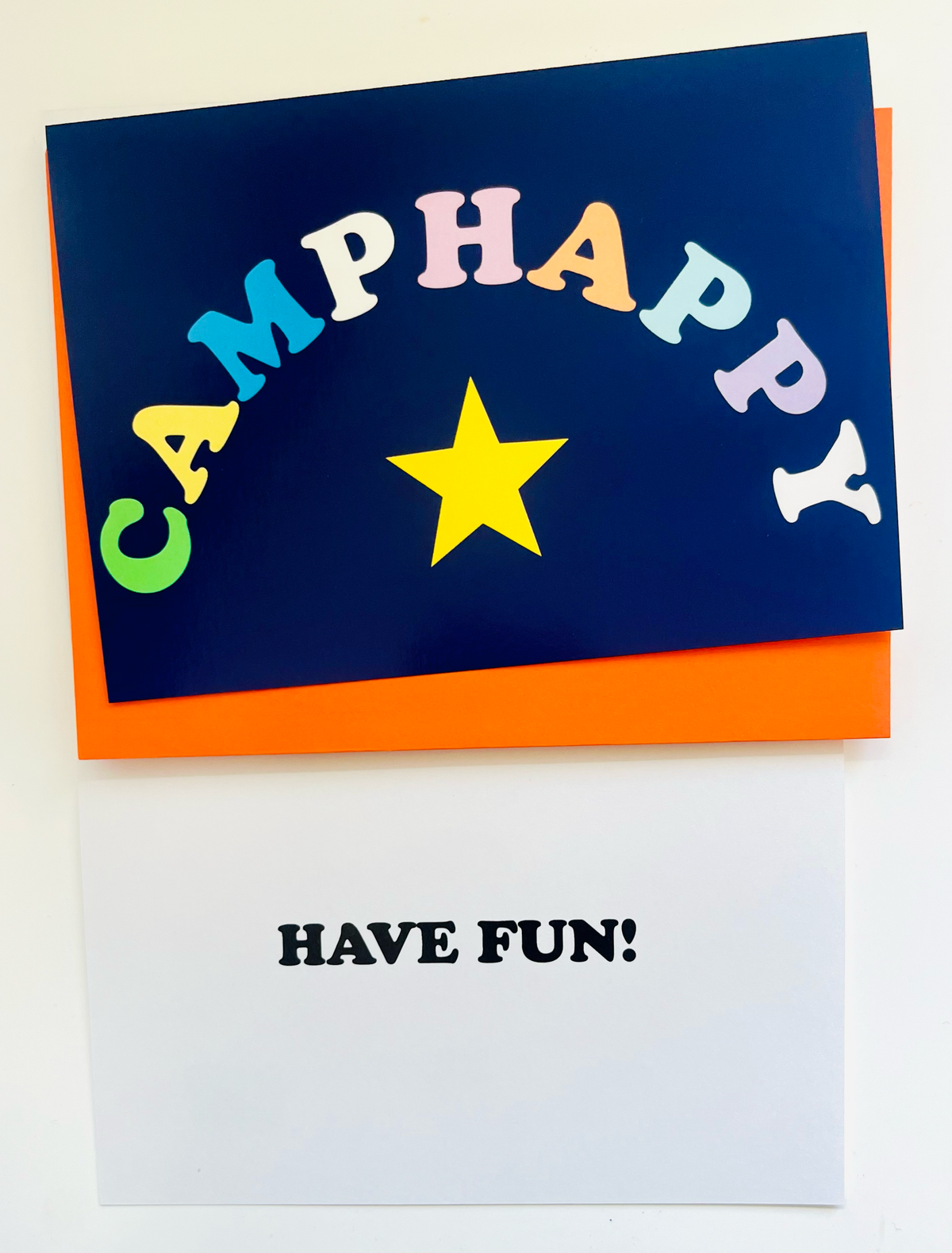 Camp Happy greeting card with colorful rainbow letters spelling CAMPHAPPY in an arch over a yellow star on navy blue background with orange envelope behind